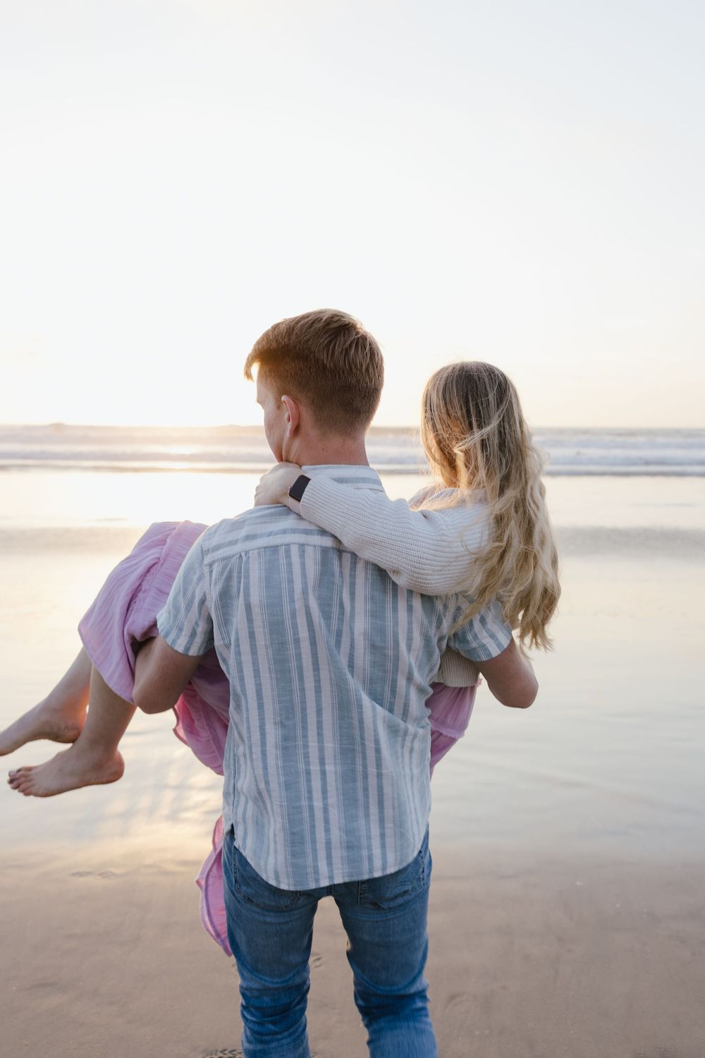 Man carrying woman in his arms on a beach at sunset, viewed from behind.