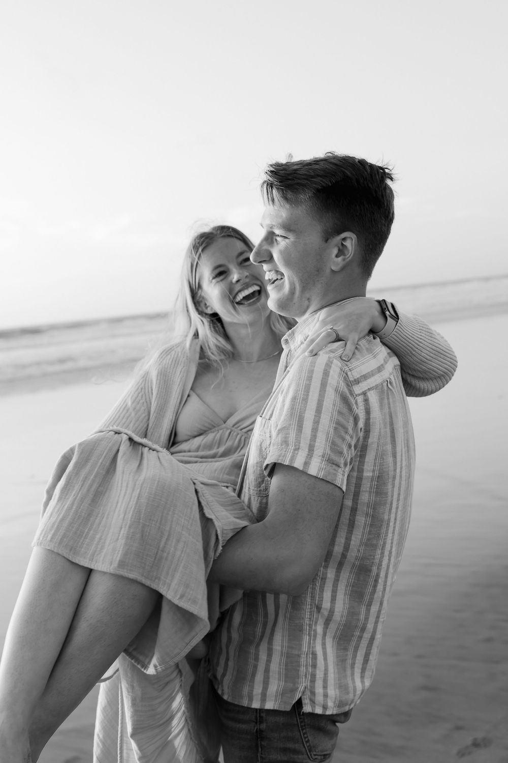 Black and white photo of couple laughing as man carries woman on a beach.