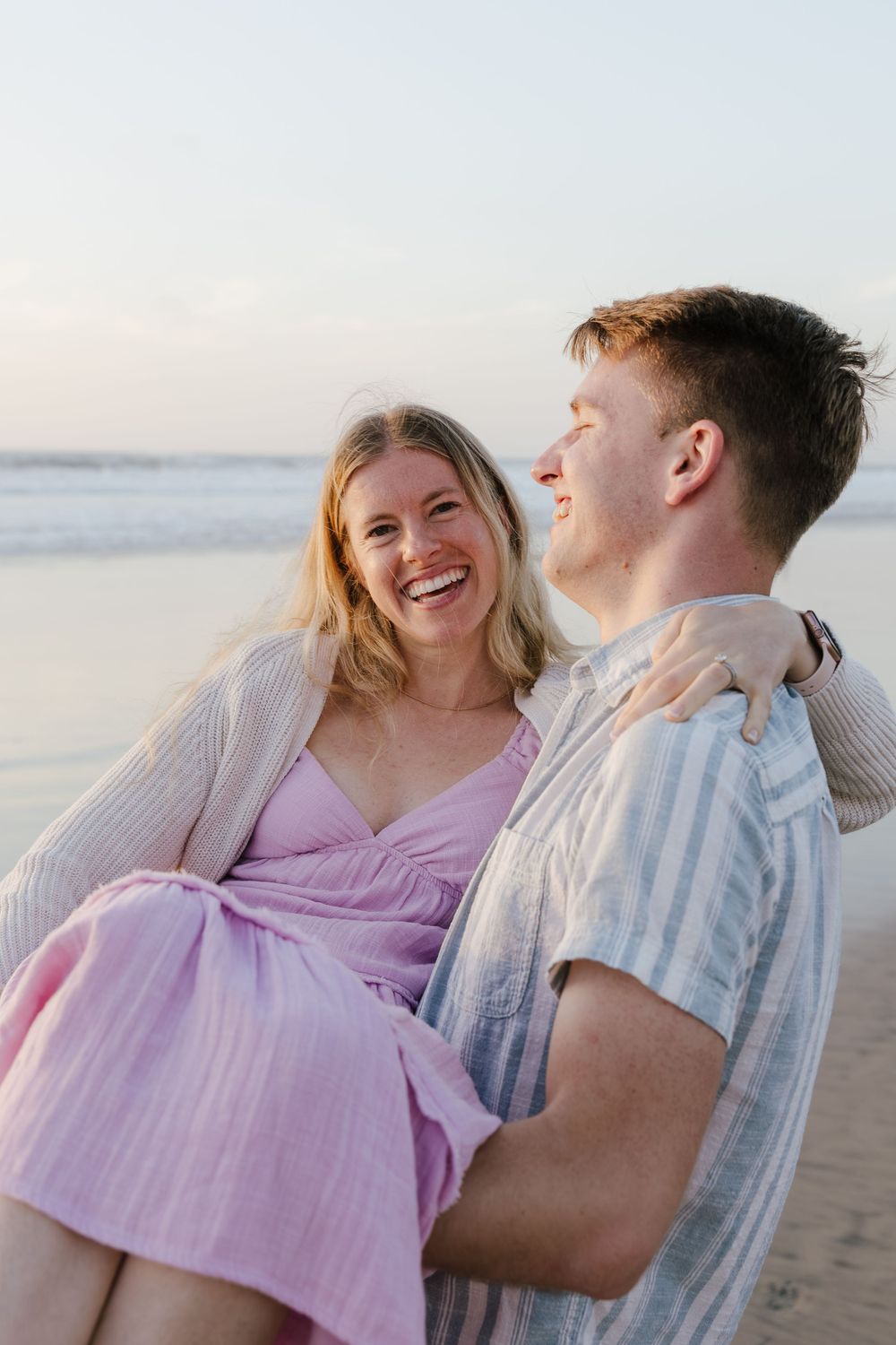 Man holding laughing woman in his arms on a sandy beach at sunset.