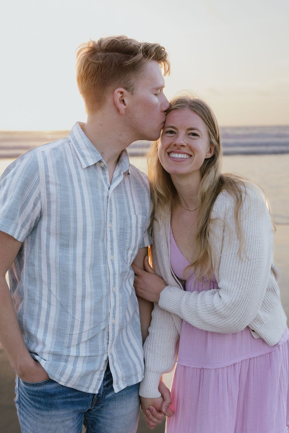 Man kissing smiling woman on the cheek while holding hands on a beach at sunset.