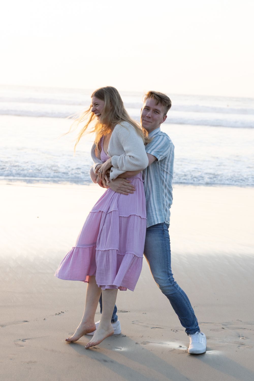Couple embracing on a sandy beach at sunset with ocean waves in the background.