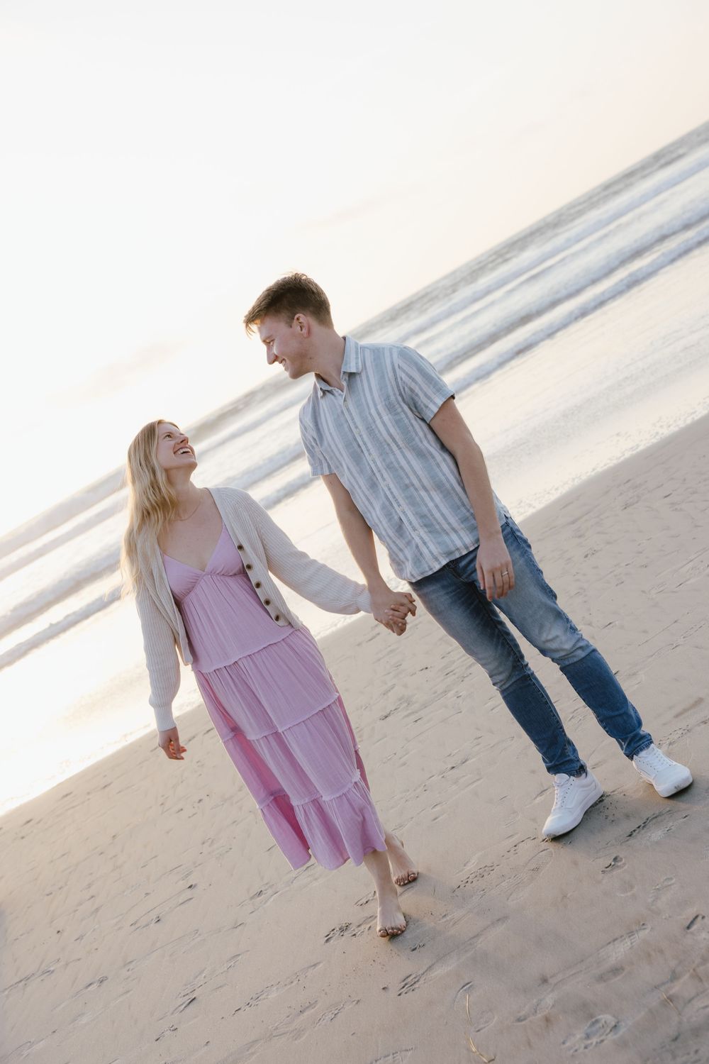 Smiling couple holds hands while walking barefoot along sandy beach at peaceful sunset.