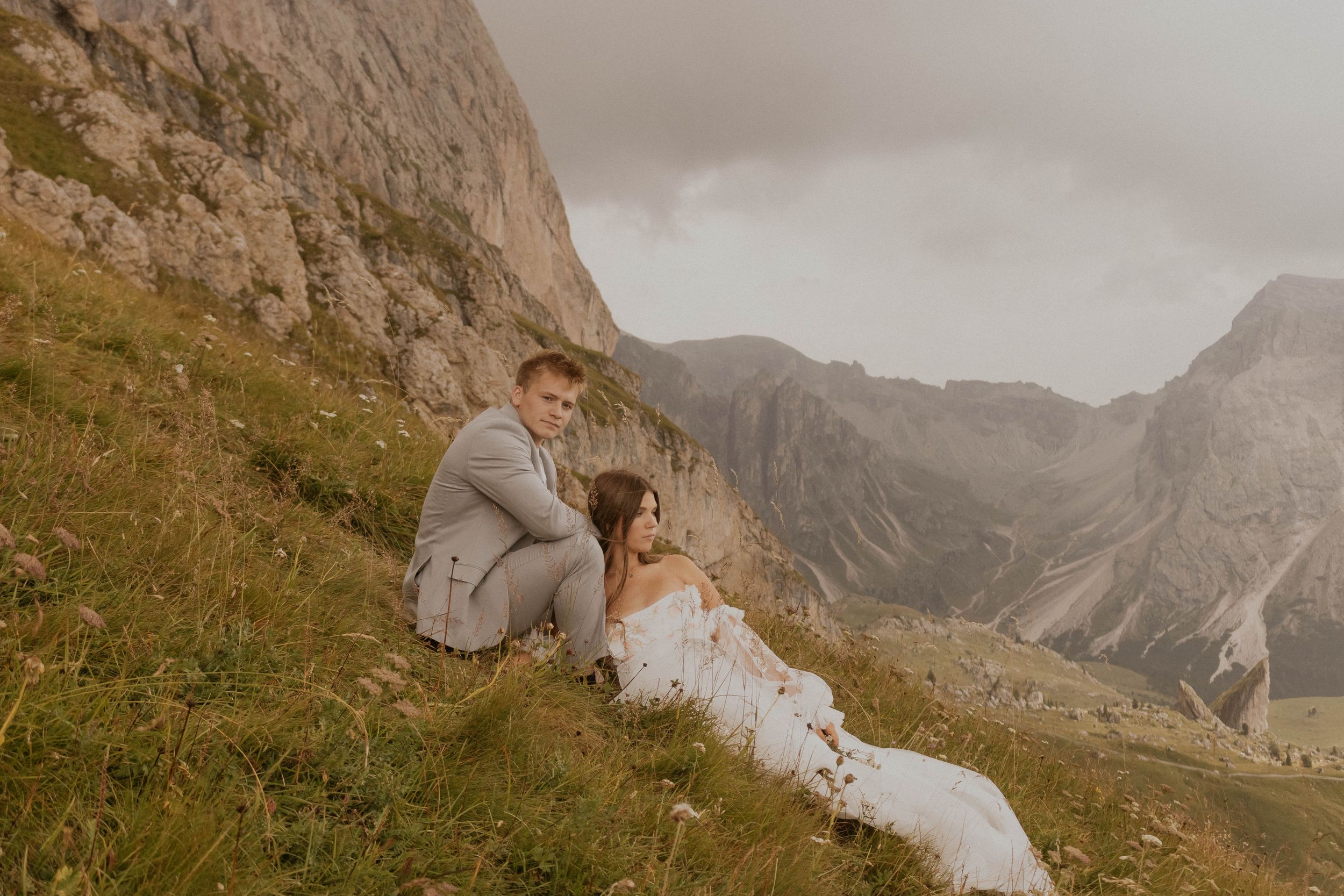A couple sits together on a mountainside overlooking a dramatic alpine valley with rugged peaks in the distance.