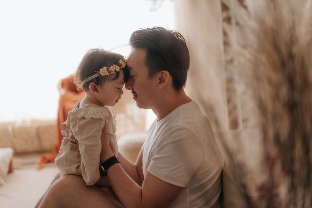 A tender moment between parent and child laying on white bedding in a warm-toned studio setting.在暖色調的攝影棚環境中，父母和孩子躺在白色床單上，享受溫馨的時刻。