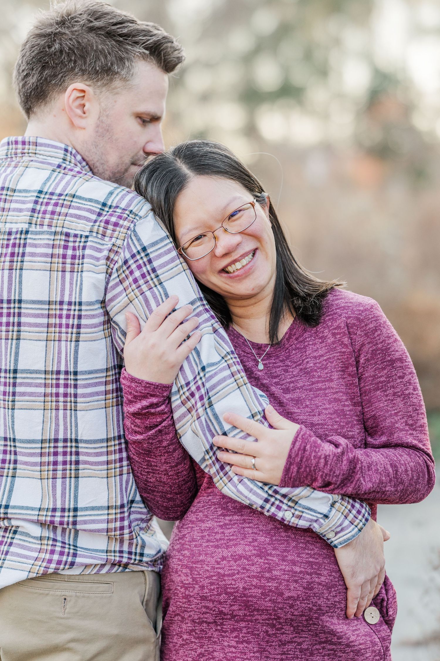 A couple embraces outdoors while wearing casual clothing, with one in a plaid shirt and the other in a burgundy sweater.