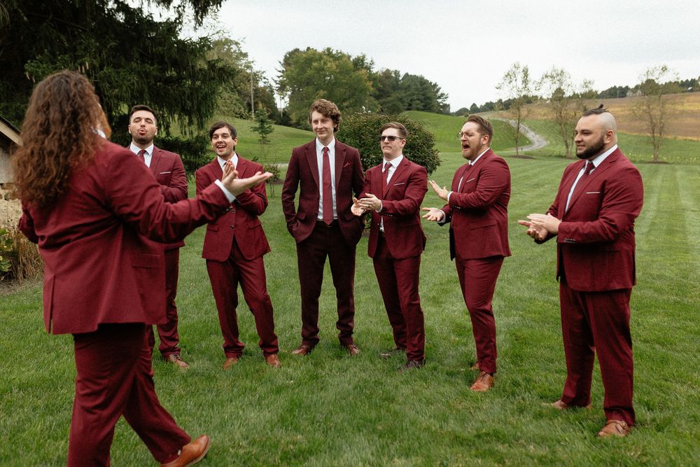 Groomsmen in burgundy suits gather in a circle on a grassy hillside.