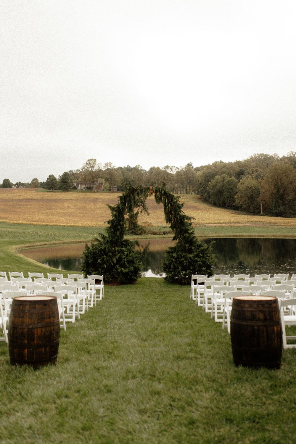 Outdoor wedding ceremony setup with white chairs and rustic wine barrels flanking a greenery arch by a serene pond.