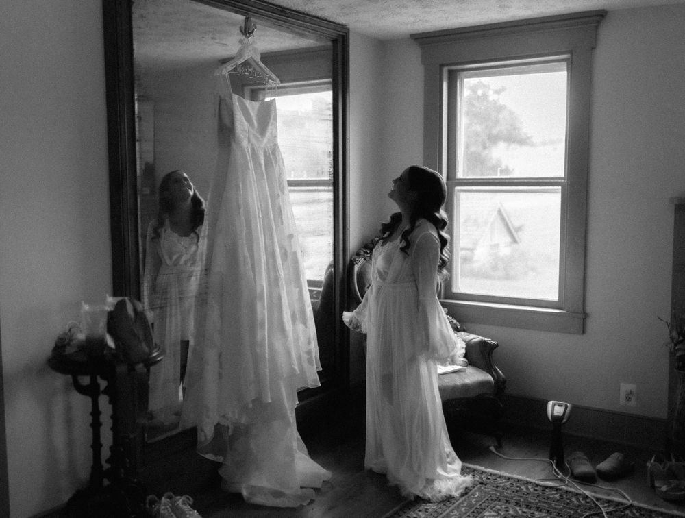 A black and white photograph shows a wedding dress hanging in a room while someone in white stands nearby.