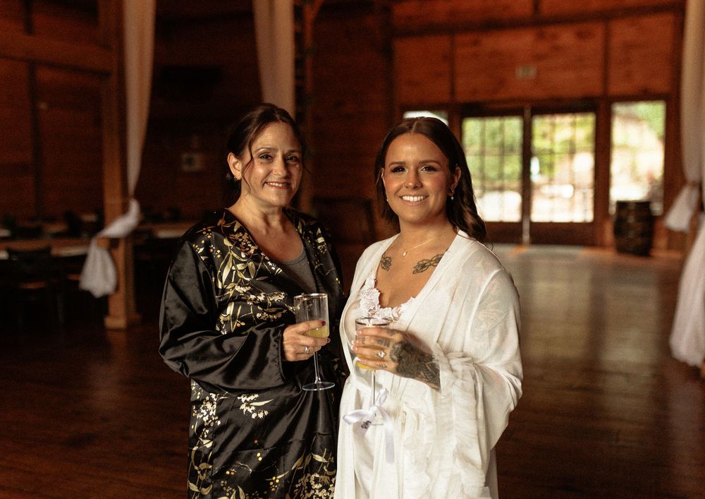 Two women in elegant evening attire hold wine glasses while standing in a rustic wooden interior space.