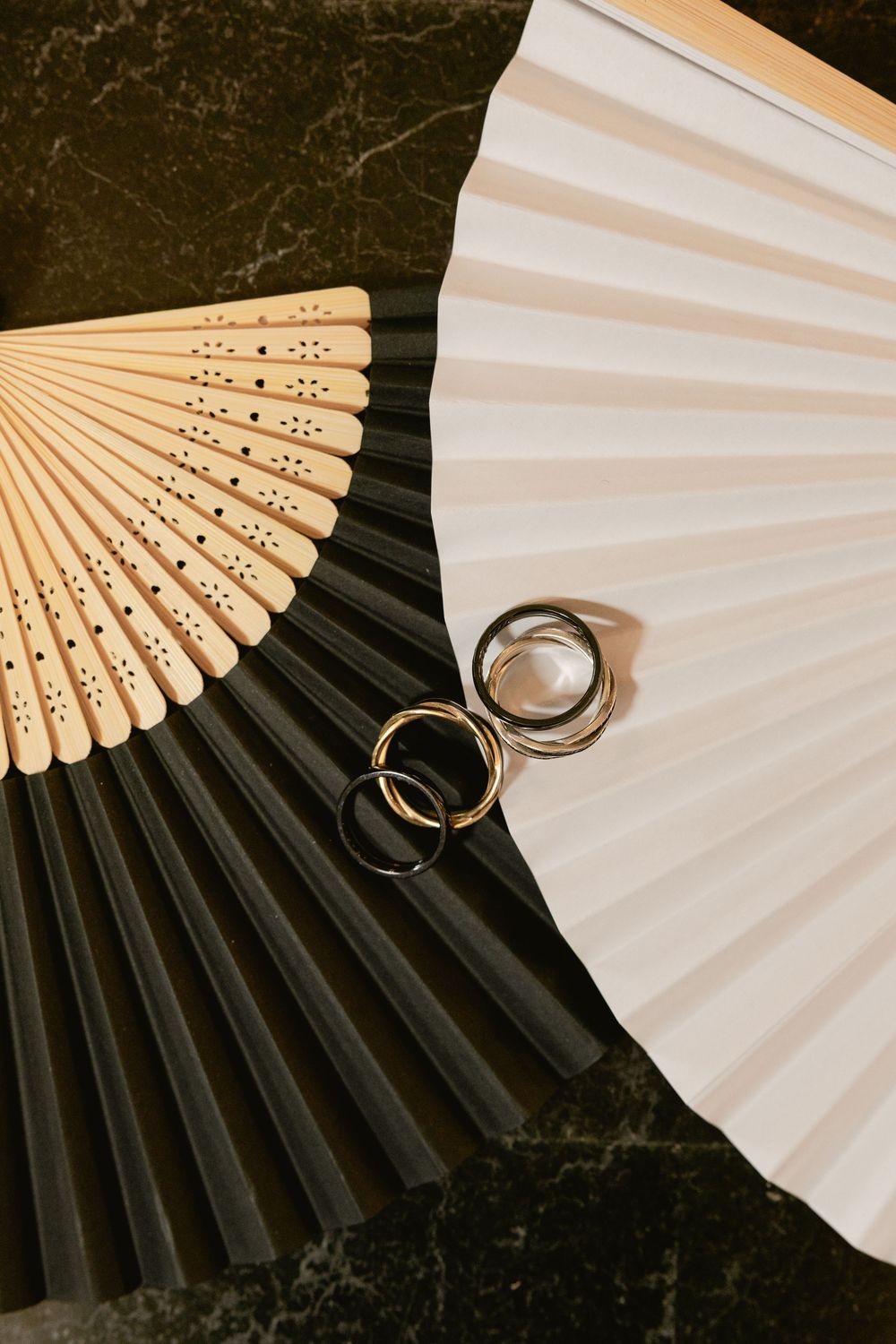 Wedding rings placed on white and black folding paper fans against dark marble background.