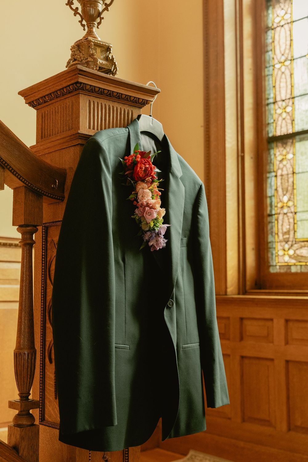 A dark green suit jacket with floral boutonniere hangs on wooden railing in church interior with stained glass windows.