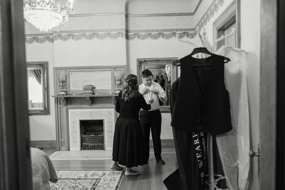 A black and white photograph shows a vintage interior room with clothing on display and people gathered near a fireplace.