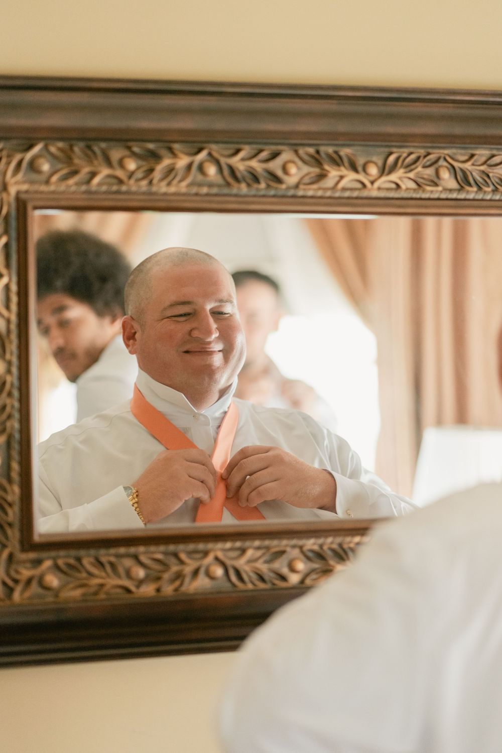 Reflection in ornate mirror shows person adjusting peach-colored tie while getting ready.