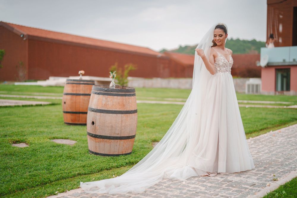 A bride in a flowing white wedding dress stands among wooden barrels outside a rustic red barn venue.