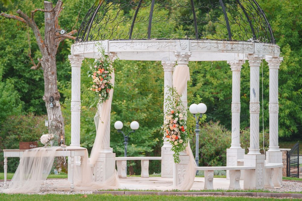 White wedding pergola decorated with floral arrangements stands in a lush green garden setting.