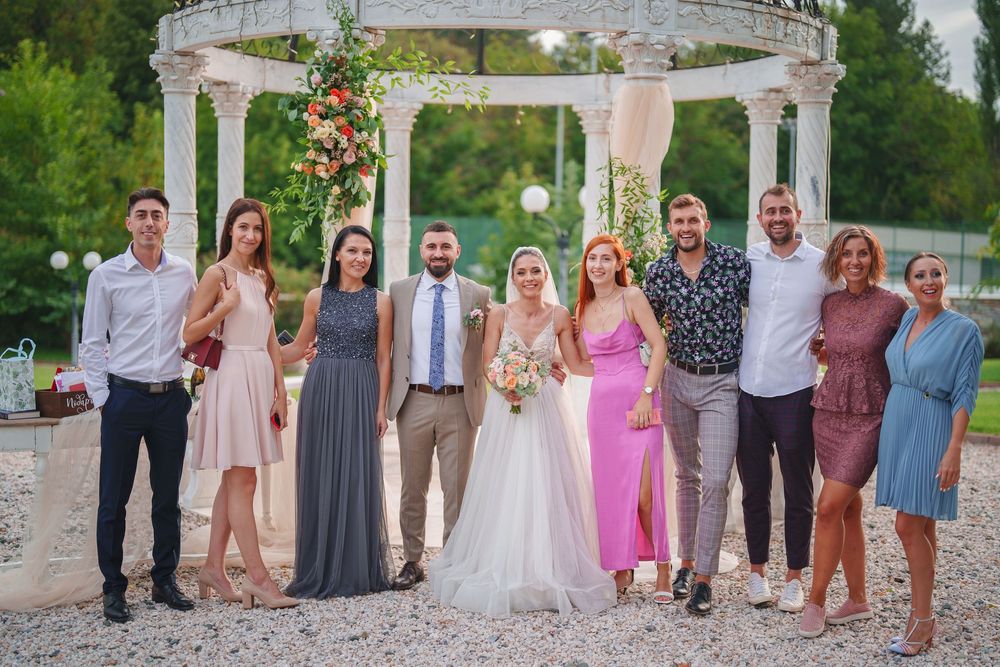 A wedding party poses together under a white floral gazebo in an outdoor garden setting.
