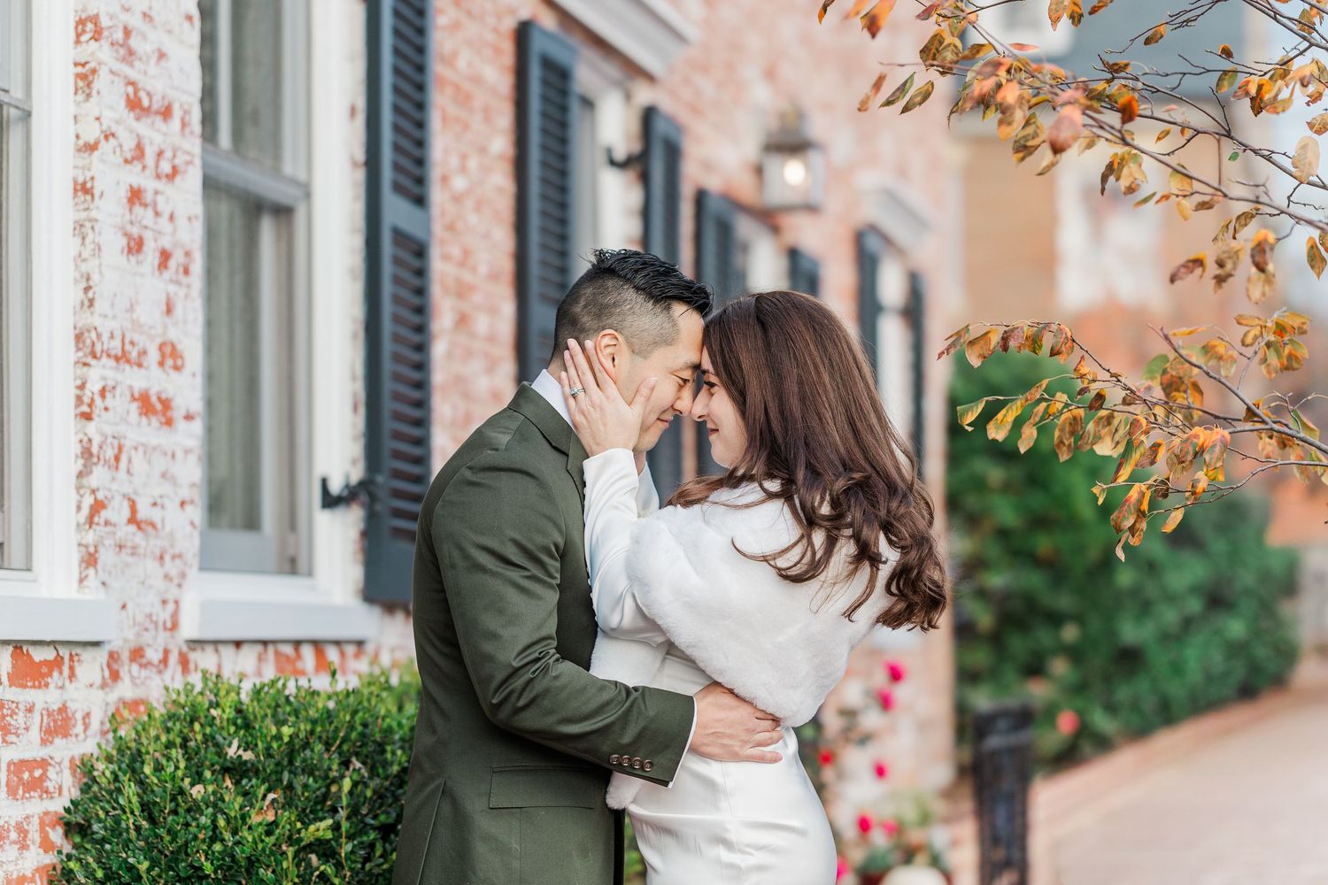 A couple shares a romantic moment in front of a brick building with autumn leaves falling around them.