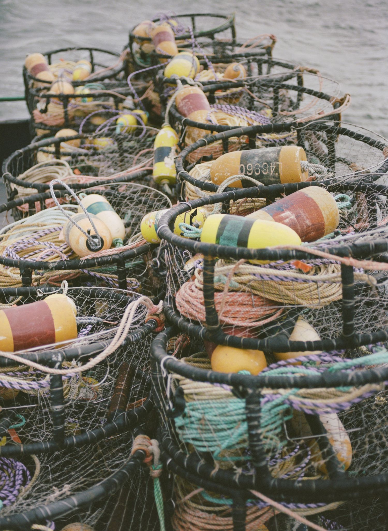 Weathered fishing crab pots with colorful buoys and ropes stacked together on a dock in muted tones.