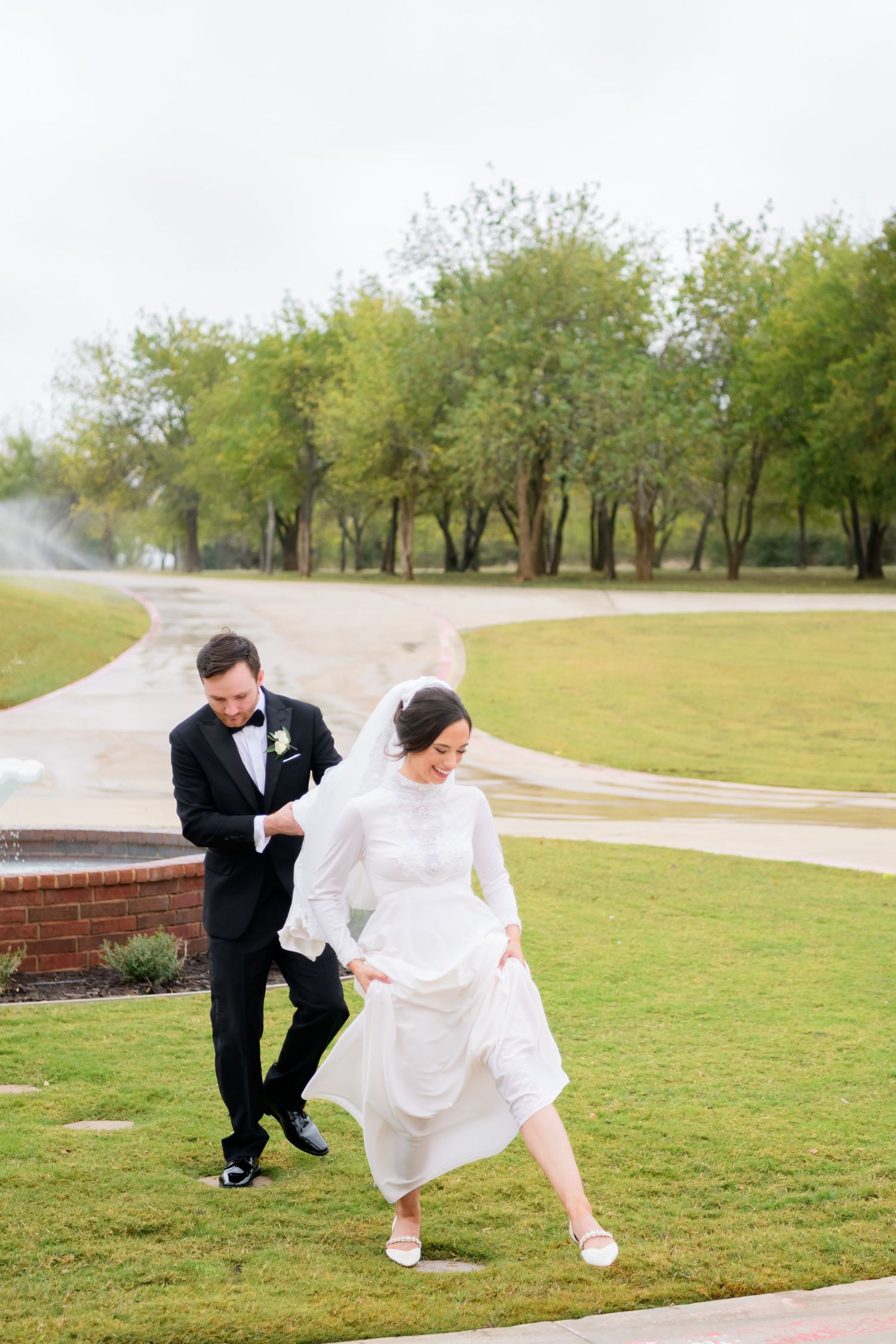 Couple in wedding attire playfully runs across a grassy hill with trees in the background on a misty day.