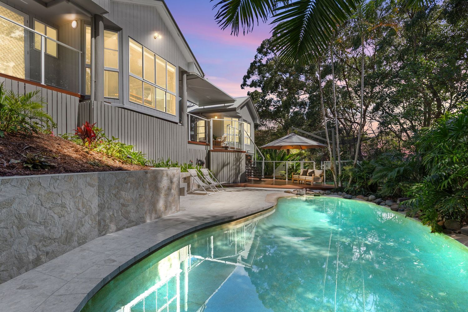 Stunning backyard pool with stone retaining walls, tropical landscaping, and outdoor entertaining area at sunset.