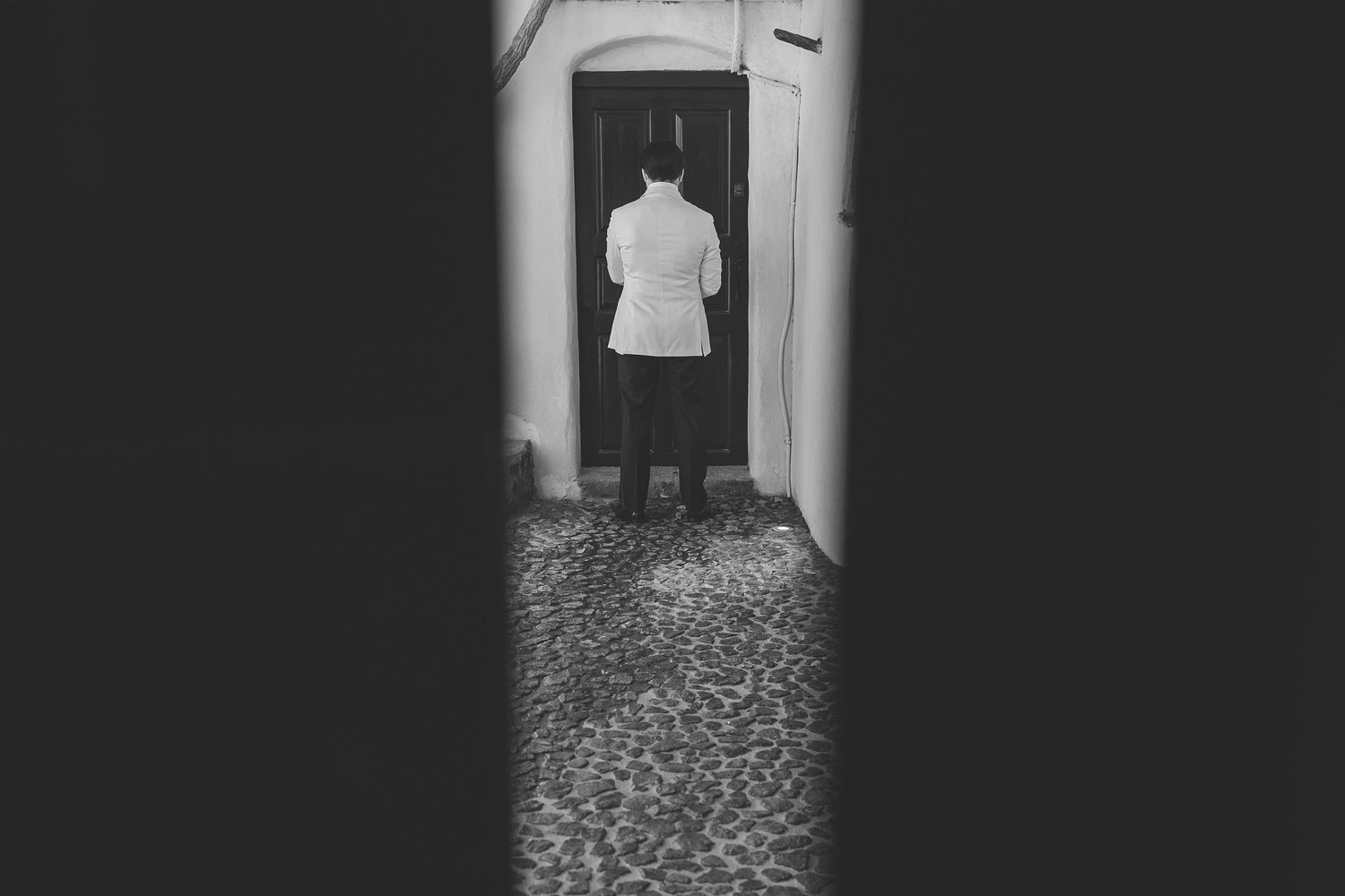 A black and white photograph shows a groom standing alone in a Santorini doorway, creating a silhouetted figure against the light.