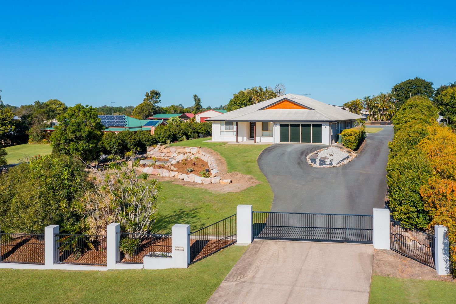 Single-story brick home with double garage, manicured lawns, and landscaped gardens behind white fenced entry in Queensland.