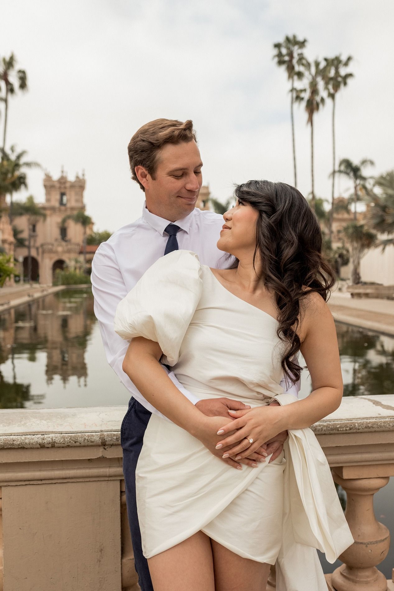 Romantic couple embraces near a reflecting pool surrounded by palm trees at Balboa Park in San Diego.