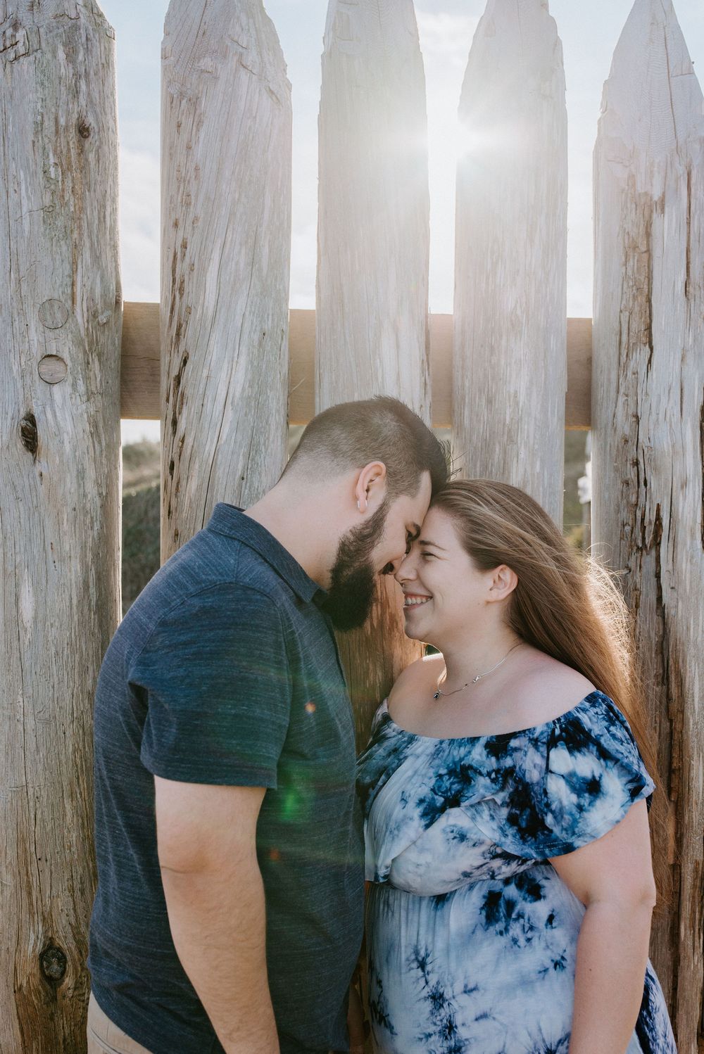 A romantic couple shares a tender moment in front of a rustic wooden fence backlit by warm sunlight.