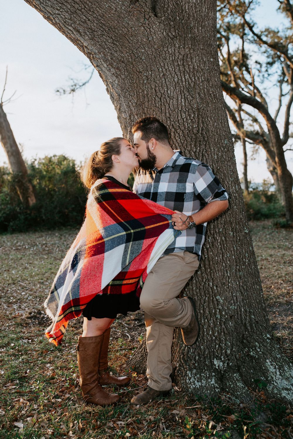 A romantic couple wrapped in a plaid blanket share tender moments against a tree trunk in an outdoor setting.