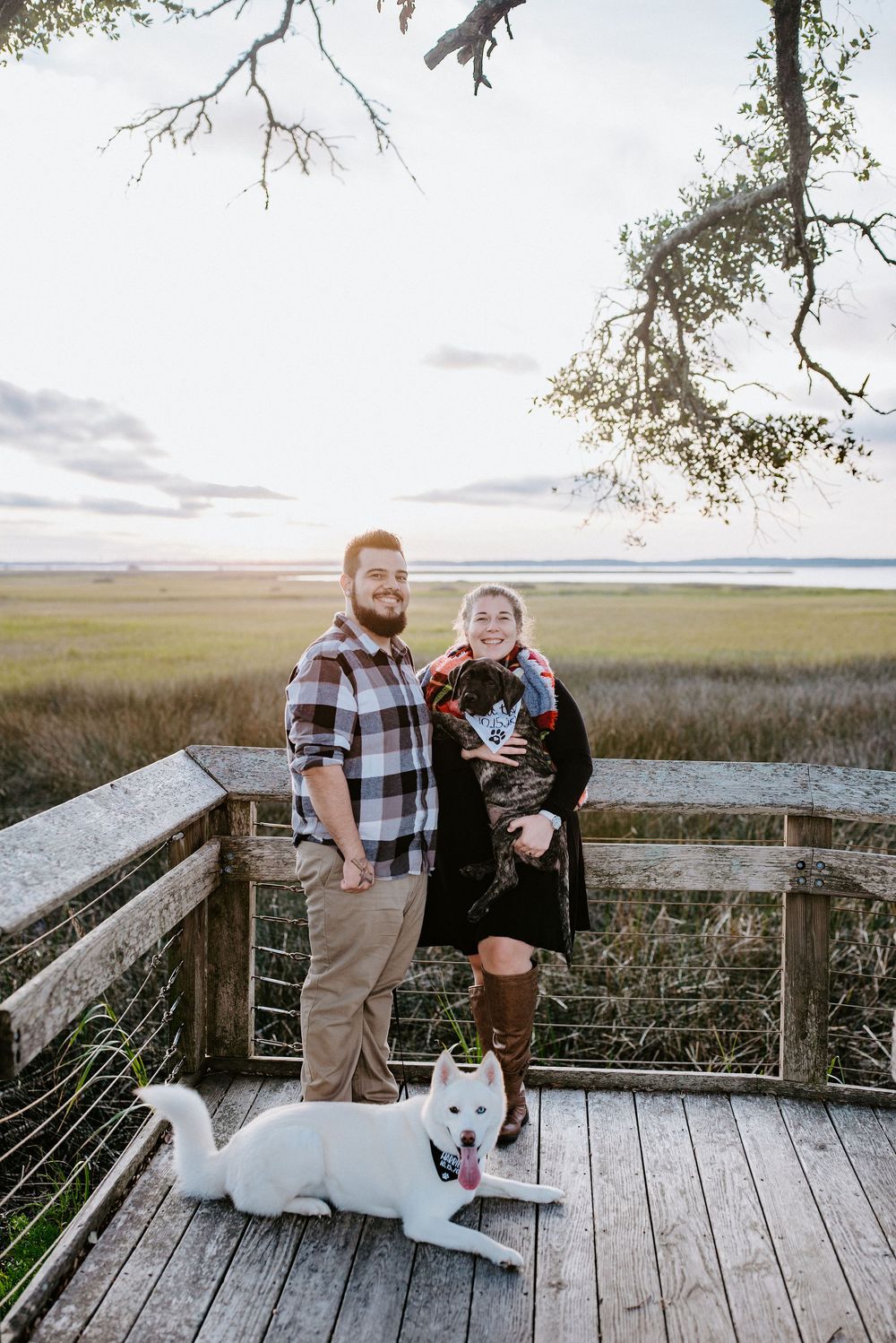 Couple and their white dog enjoy scenic views from wooden boardwalk overlooking coastal marshland at sunset.