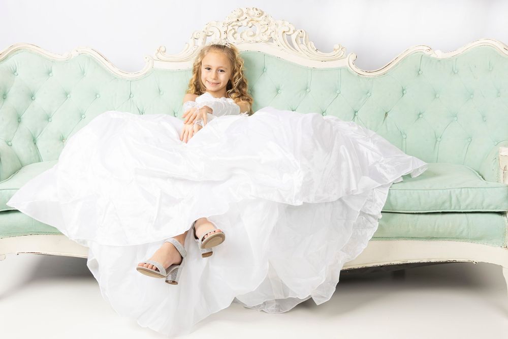 A flowing white christening gown with delicate lace details is artfully arranged on a mint green tufted sofa.