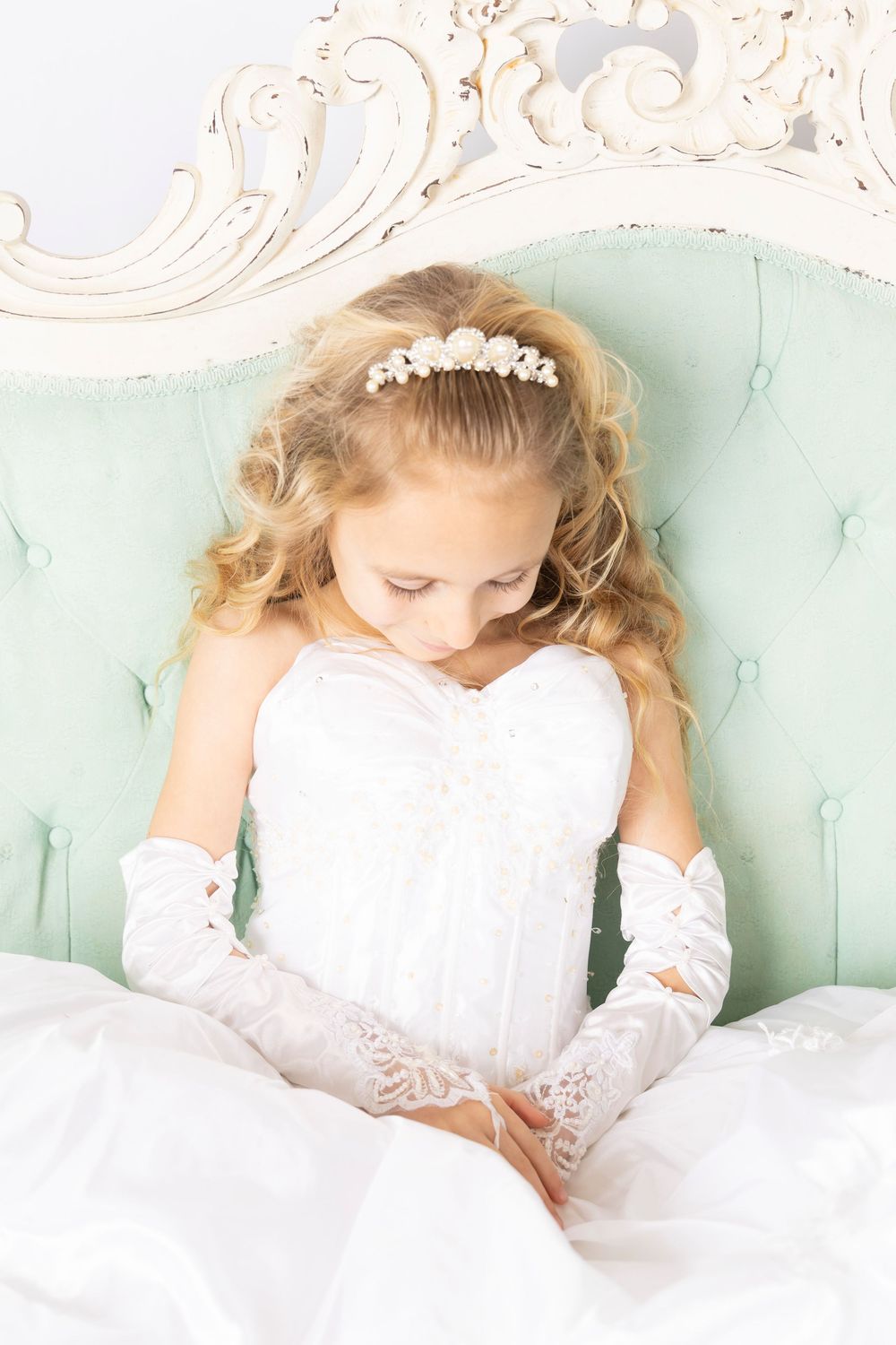 Young girl in white dress and long gloves sitting on mint green vintage chair looking down pensively.