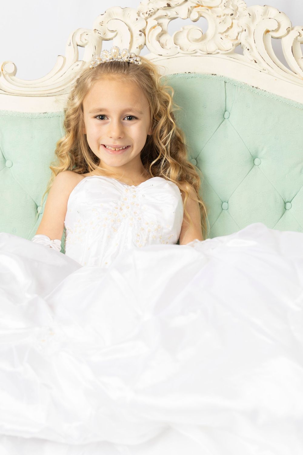 Child in white dress sitting on ornate mint green chair with decorative headpiece.