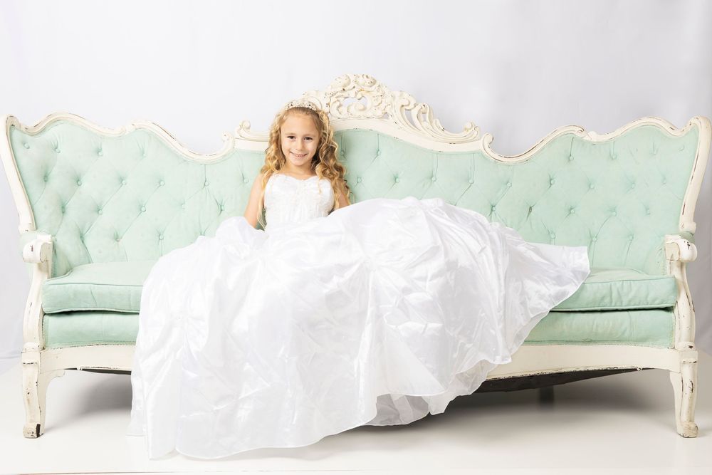 A bride in a flowing white gown reclines on a mint green vintage chaise lounge in an elegant setting.