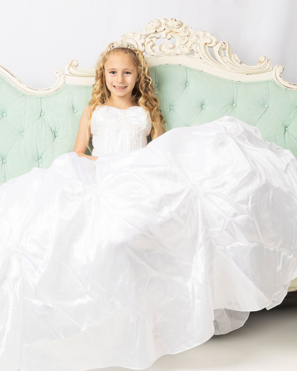 Young girl in white dress sitting elegantly on mint green vintage sofa with ornate details.