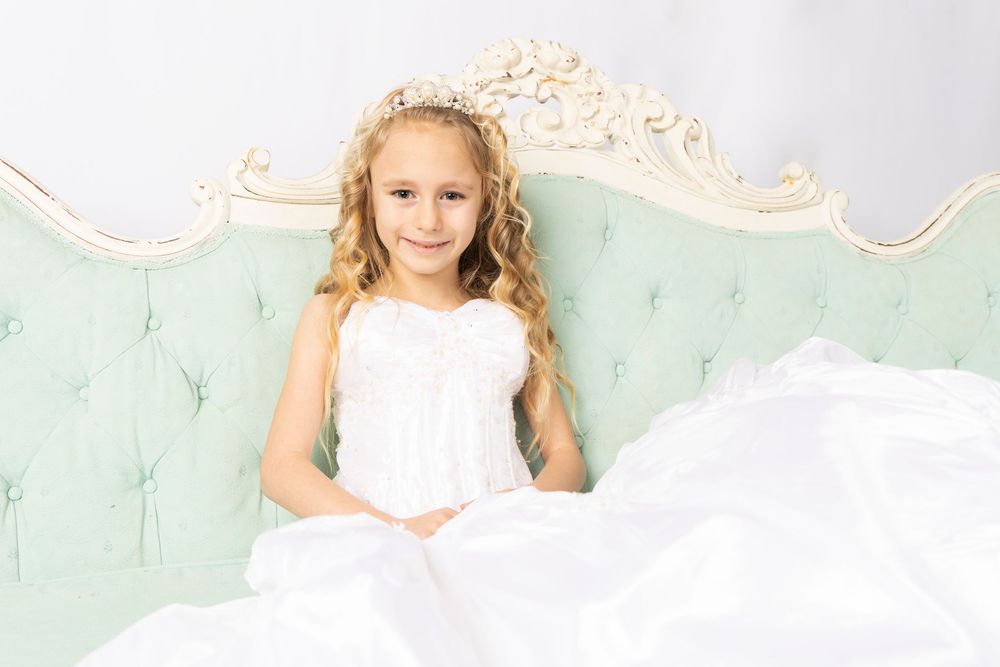 Child wearing flowing white dress poses gracefully on antique mint sofa.
