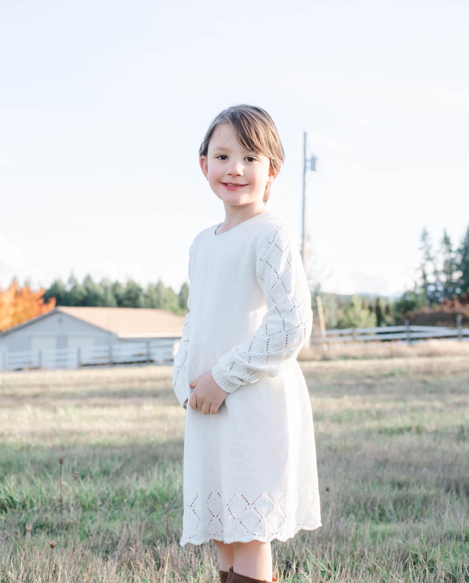 Child in white lace dress plays in grassy field at sunset with rural buildings in background.