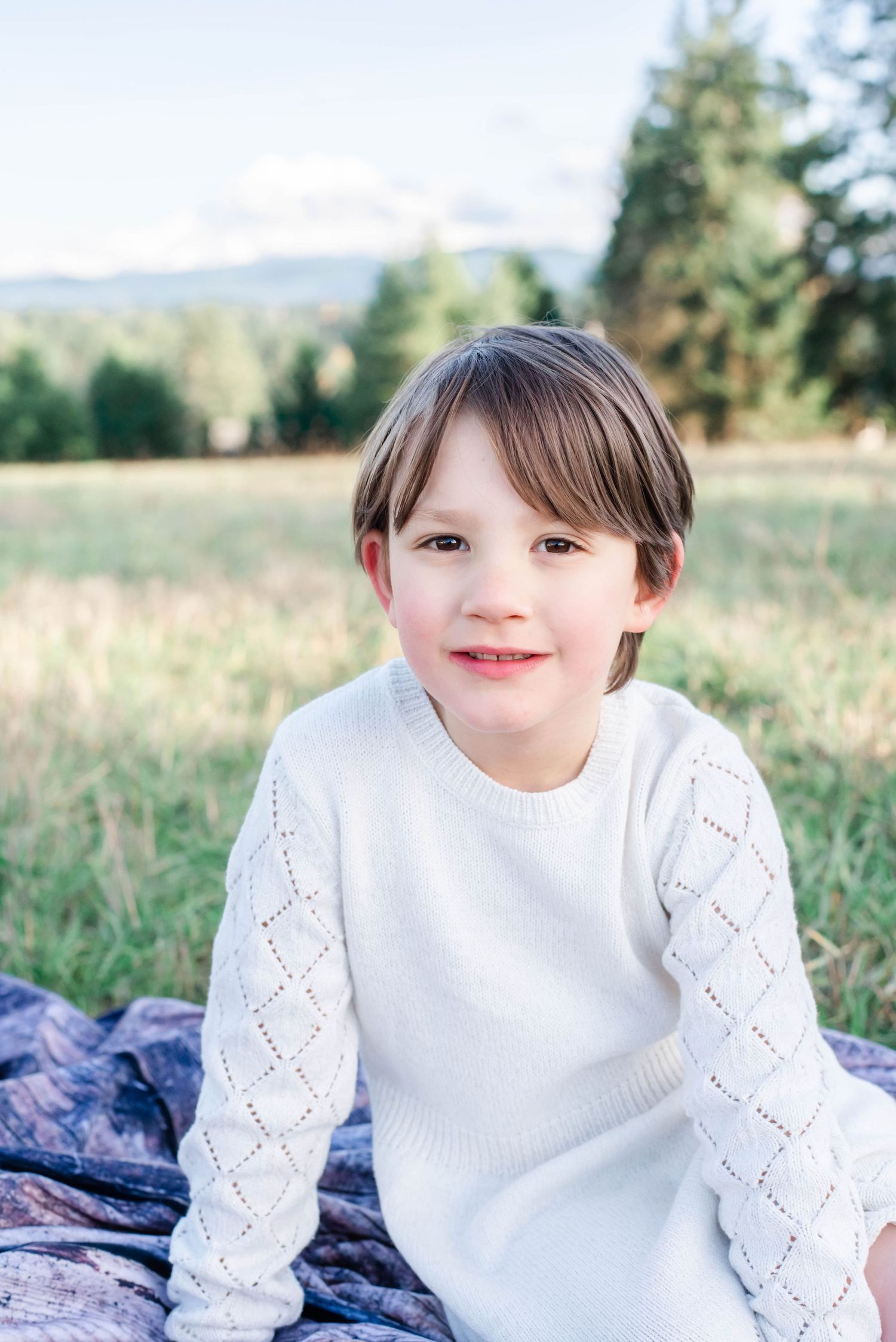 A child in a white sweater sits outdoors on a blanket in a grassy field on a sunny day.