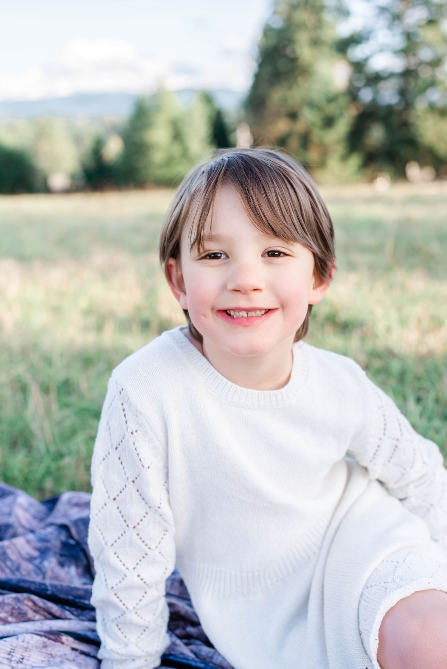 Child in white eyelet dress sits outdoors on blue blanket with trees and mountains in background.