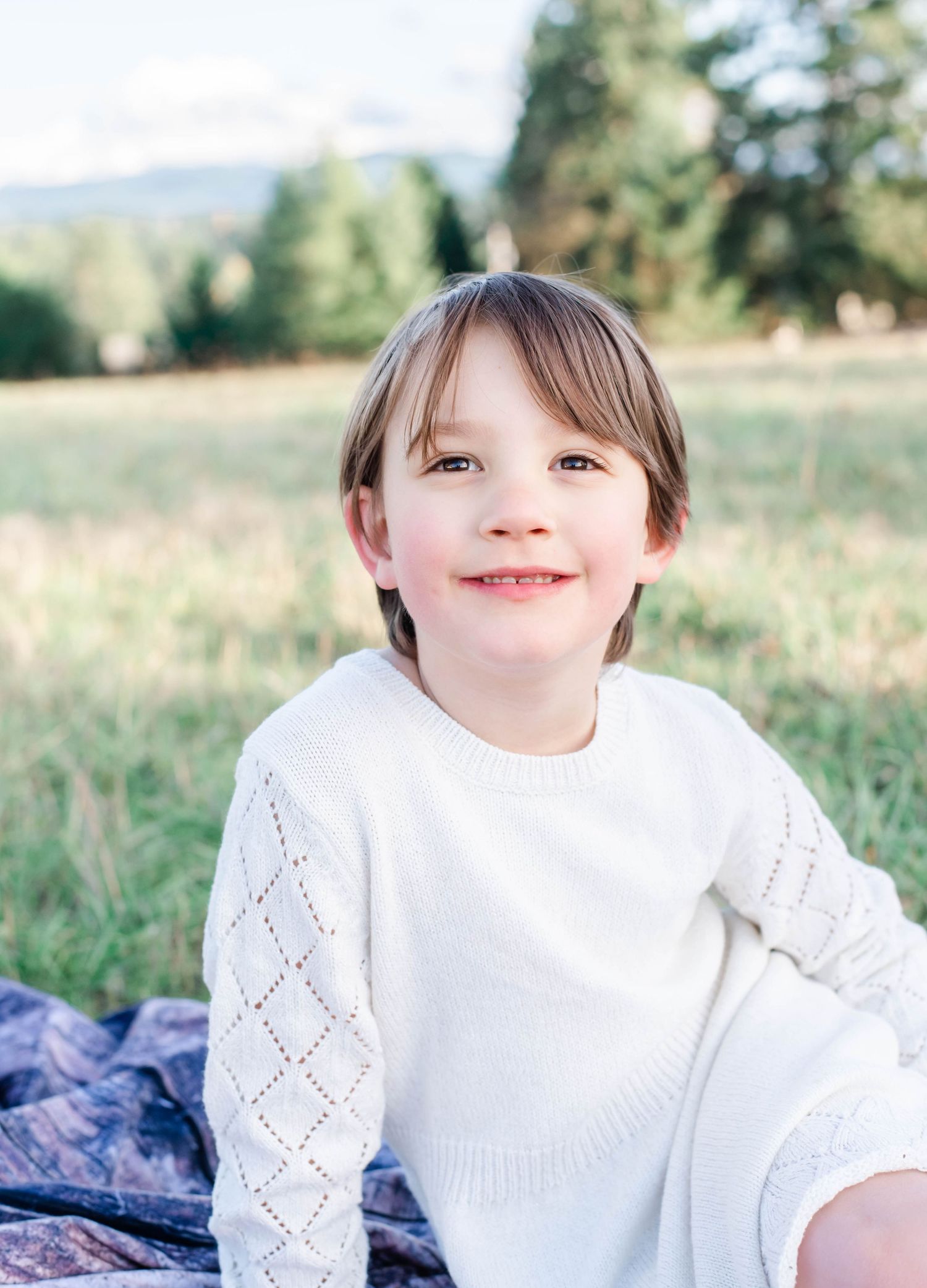 Child in white sweater smiling outdoors in natural setting with blurred background.