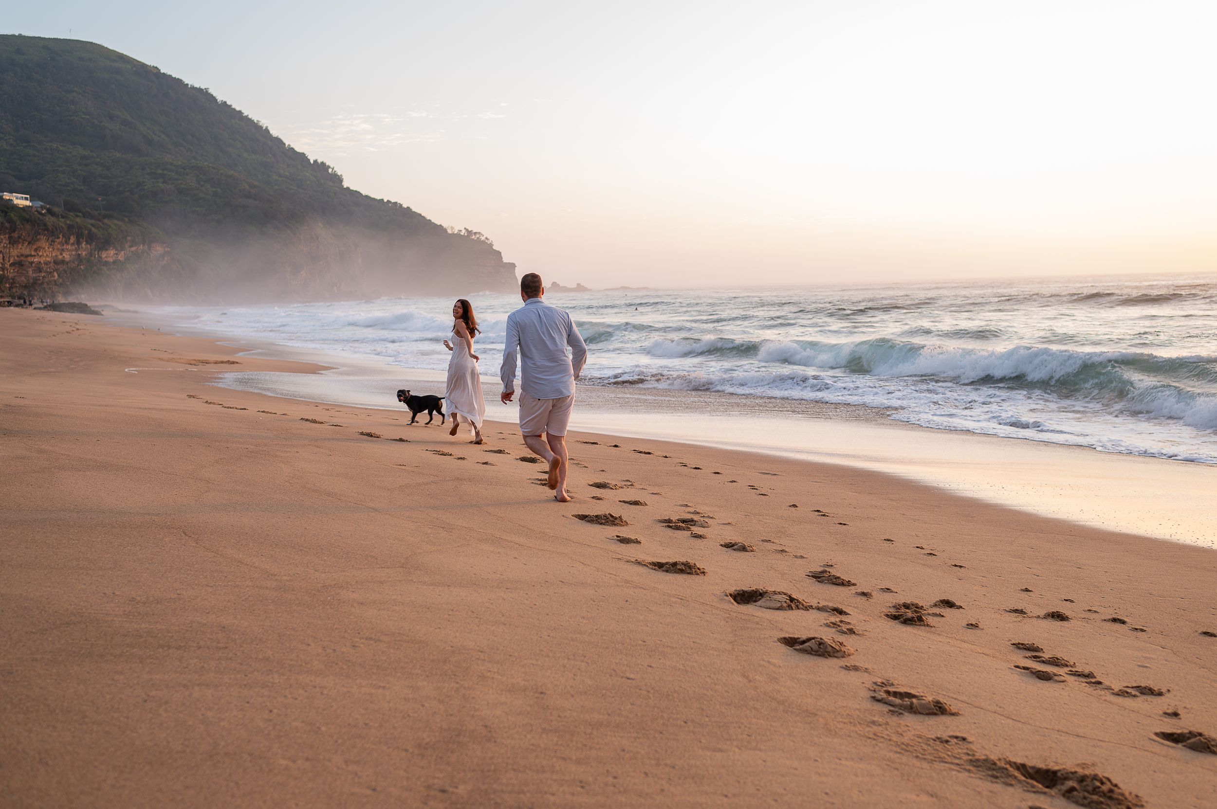 A couple walks along the sandy beach at sunset with their black dog following behind them.
