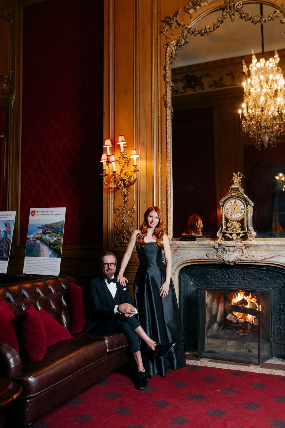 Elegant couple poses in formal attire by ornate fireplace with chandelier in luxurious vintage room with red decor.