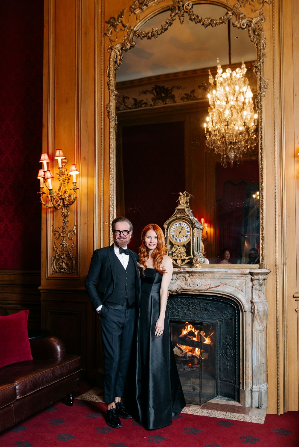 Elegant couple in formal evening wear poses by ornate fireplace with crystal chandelier in luxurious vintage room.