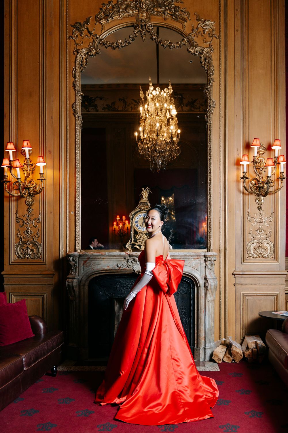 Elegant red ballgown photographed in opulent room with ornate mirror, chandelier and wall sconces against wood paneling.
