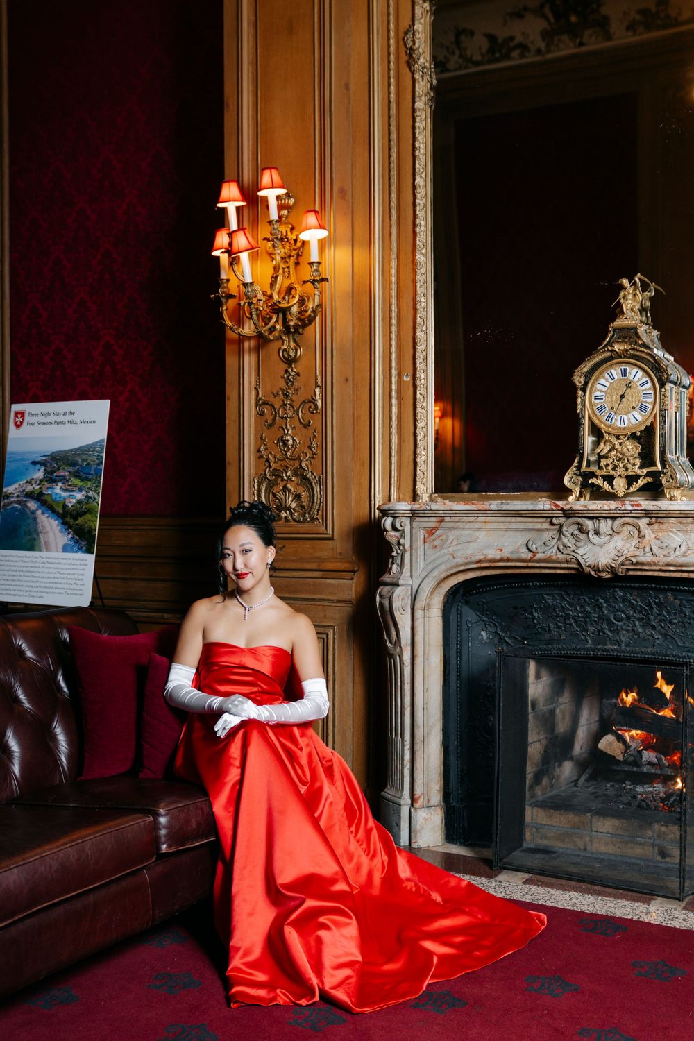 A stunning red evening gown glows against rich burgundy walls in an ornate room with a fireplace and golden details.