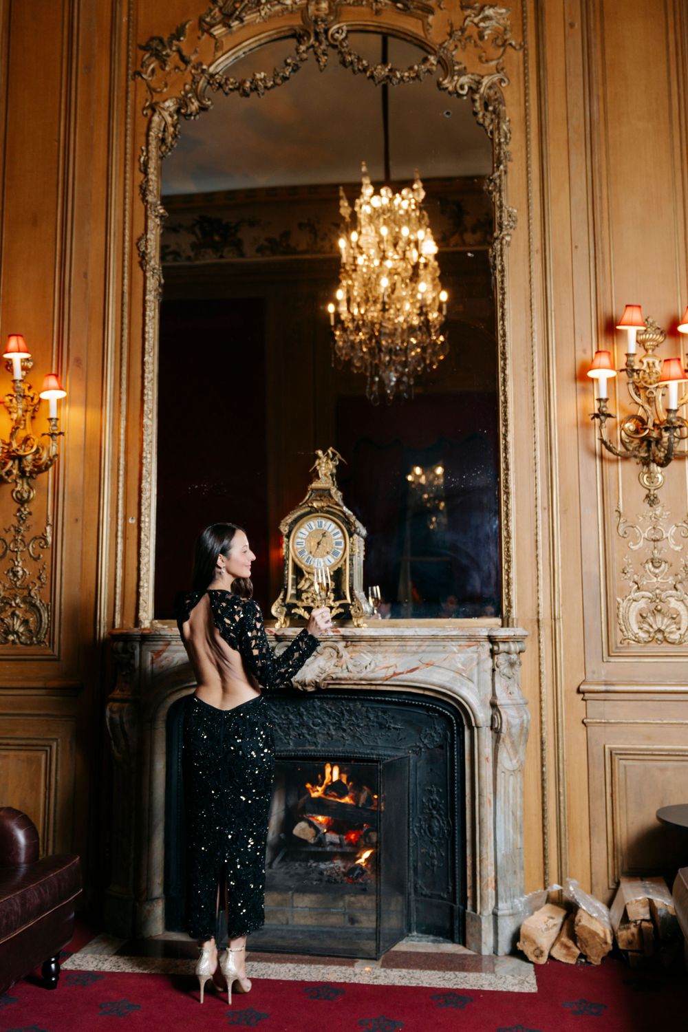Elegant black sequin gown sparkles by ornate fireplace with crystal chandelier in luxurious vintage room.
