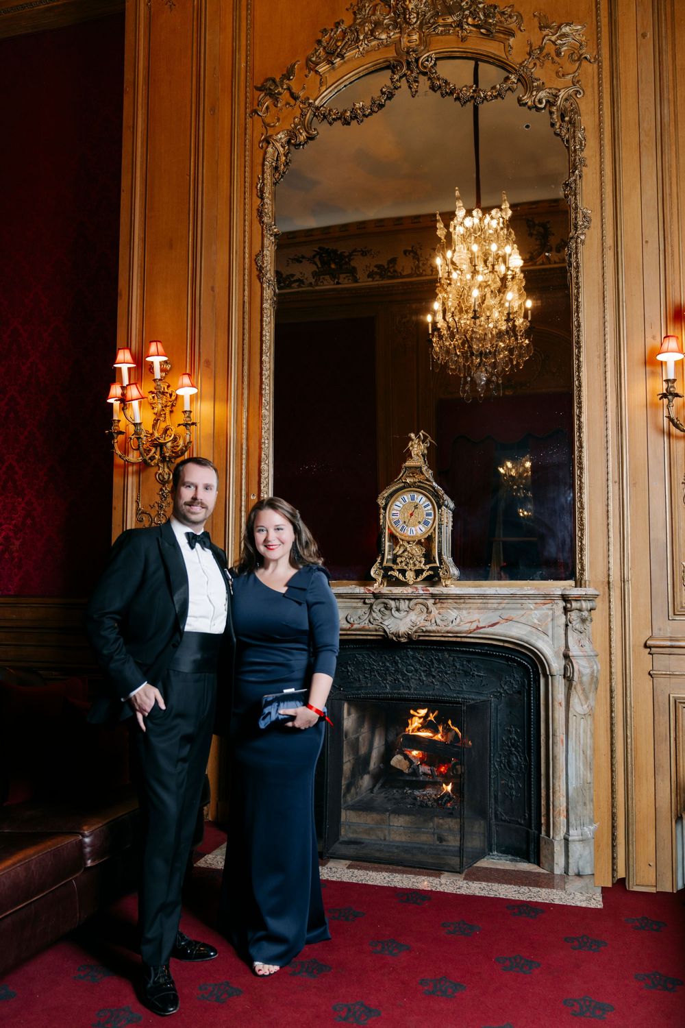Couple in formal attire poses elegantly in front of an ornate fireplace with chandeliers and red carpet in a luxurious room.