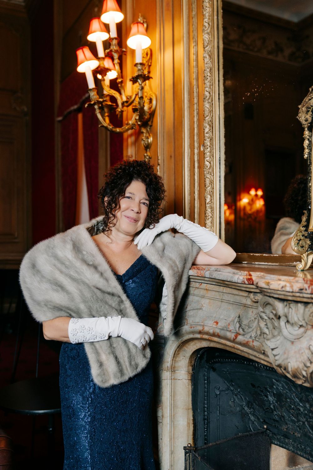Elegant evening scene with navy blue gown and grey fur wrap against ornate fireplace with vintage lighting fixtures.