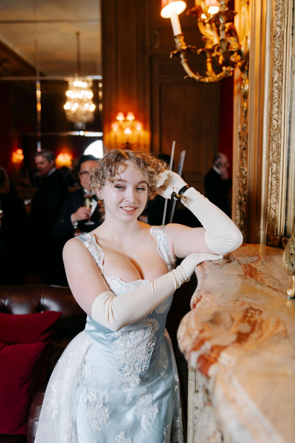 A person in a light blue formal dress poses elegantly in an ornate room with chandeliers and gold-framed mirrors.