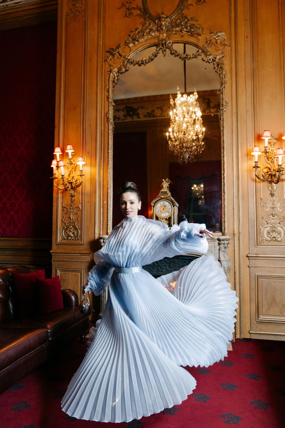 A figure in a flowing light blue pleated gown twirls dramatically in an ornate palace room with chandeliers and wall sconces.
